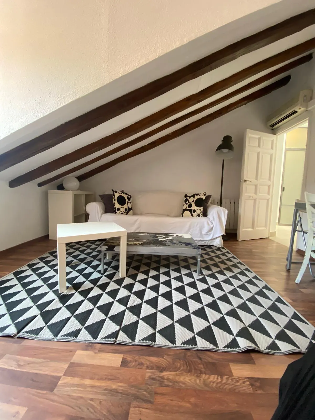 Attic living room with a white sofa, geometric rug, and wooden ceiling beams over hardwood floors.