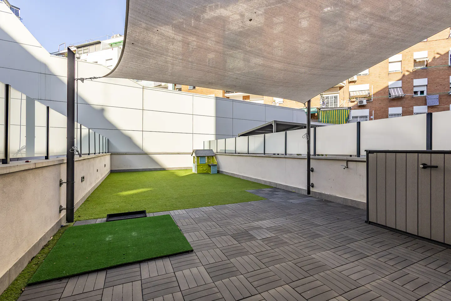 Outdoor patio with gray pavers, artificial grass, and a small playhouse. A shade sail provides cover.