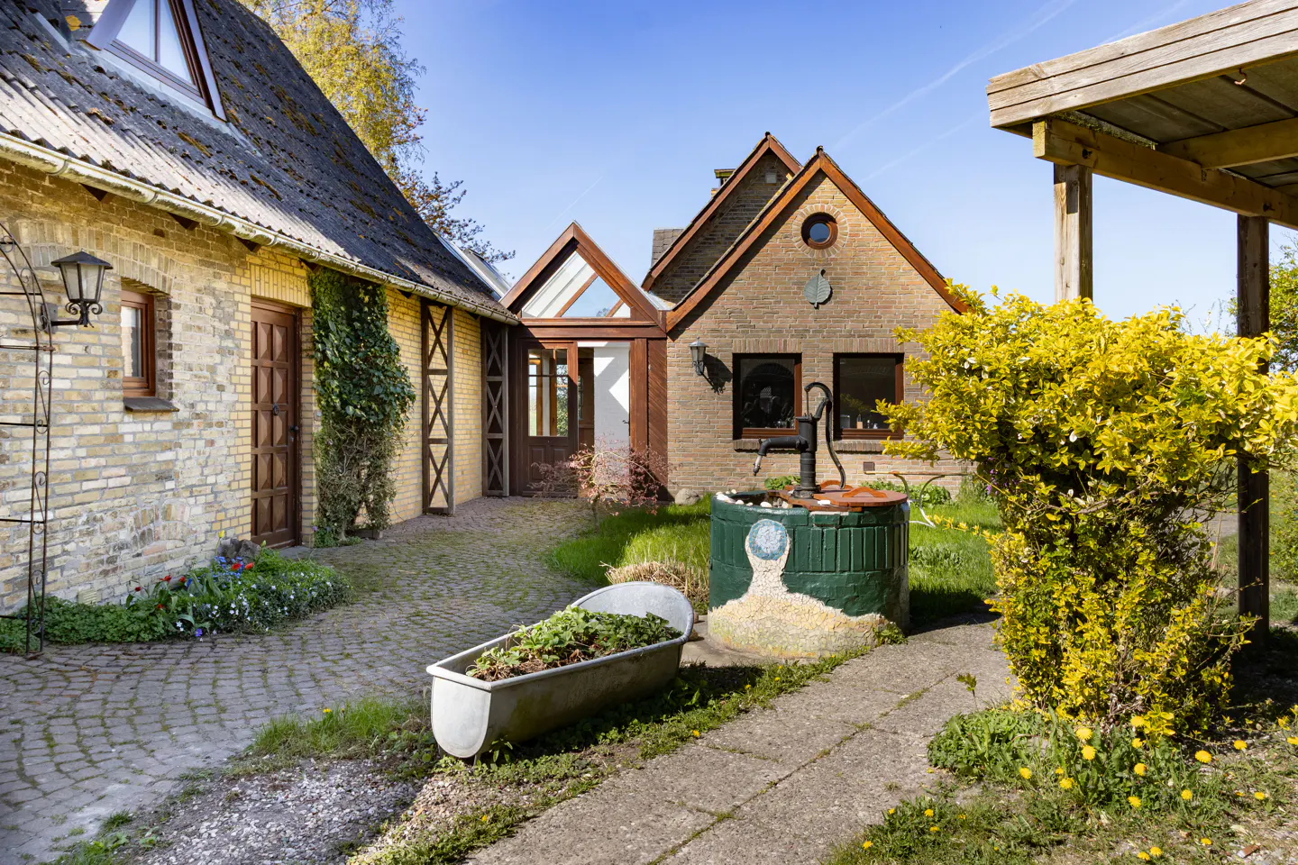 Exterior view of a brick house with a well and a trough planter on a stone path.