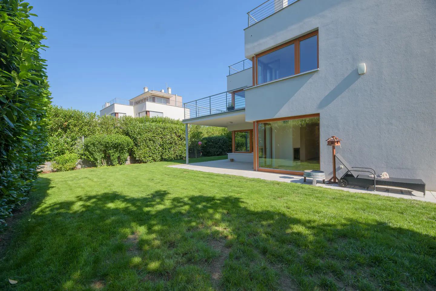 Backyard of a modern white house with a green lawn, patio, lounge chair, and trimmed hedges under a clear blue sky.