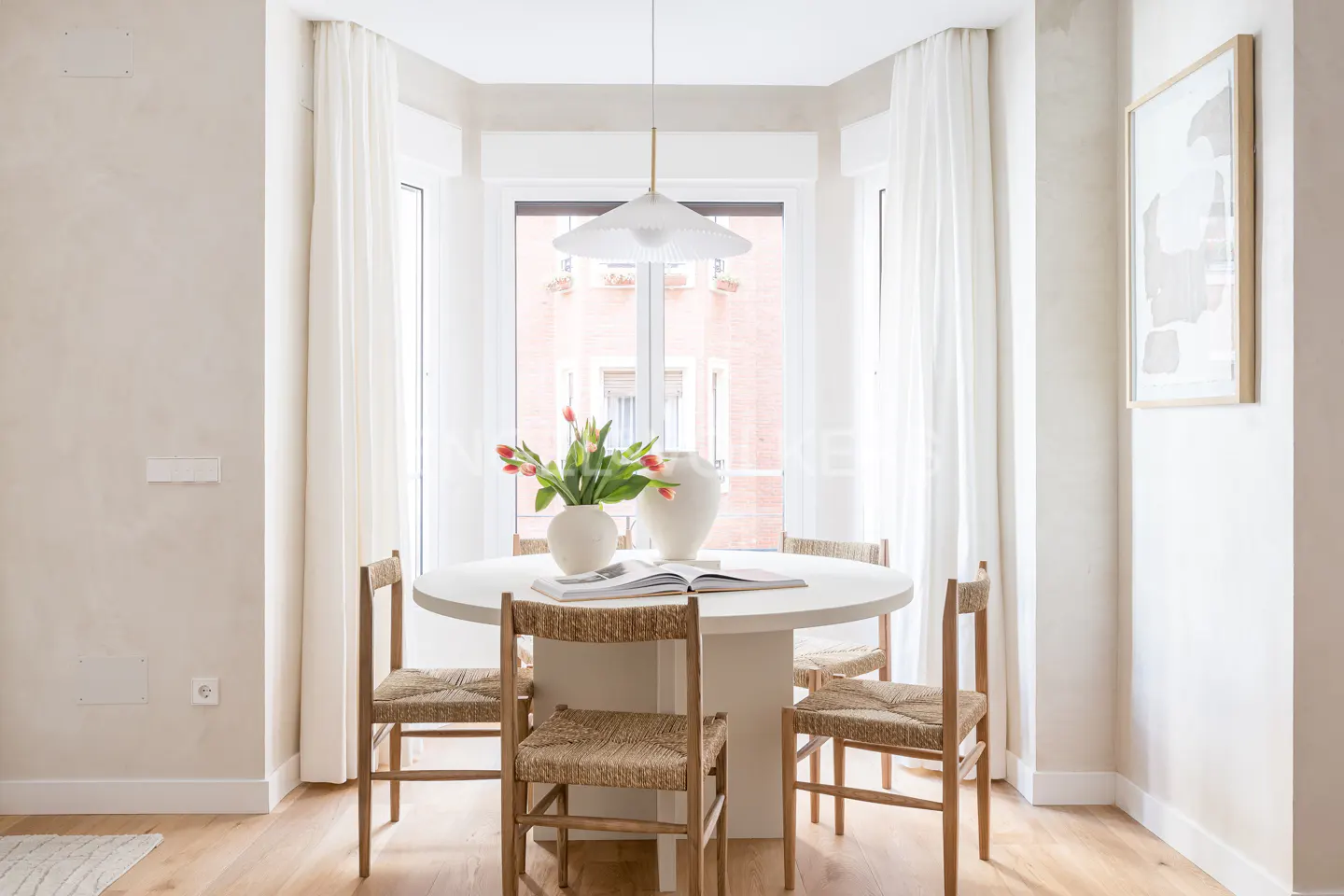 A bright dining nook with a round white table, four woven chairs, a vase of tulips, and a modern pendant light.