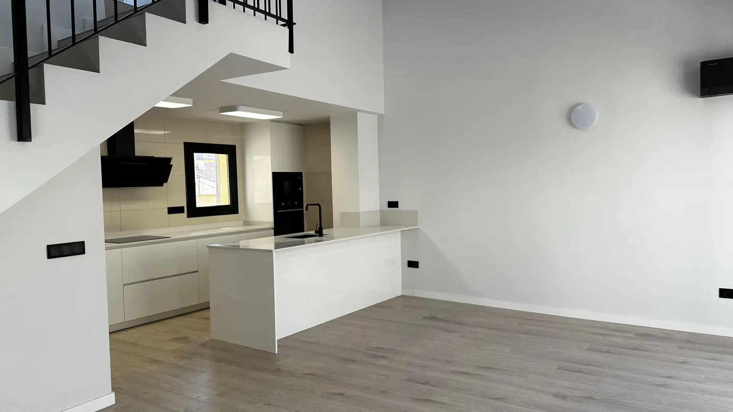 Bright, modern kitchen with white cabinets, black appliances, and light wood floors. Stairs with black railing are visible.
