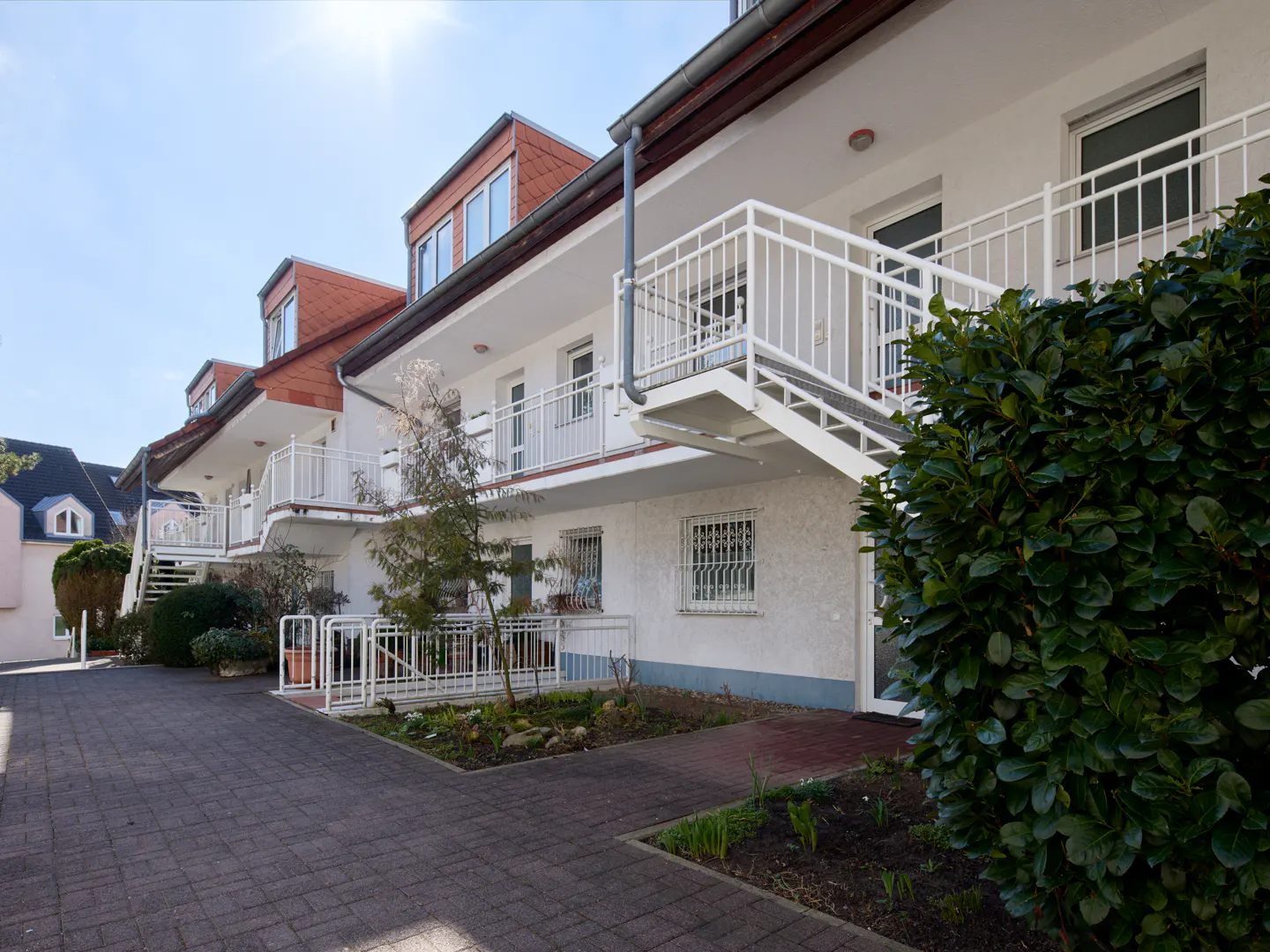 Exterior view of a white apartment building with red roofs, white railings, and a brick walkway.