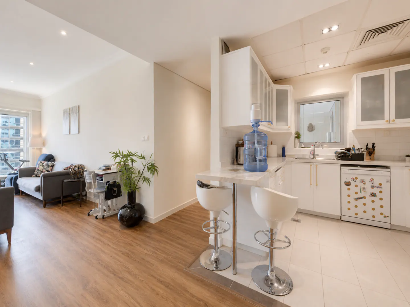 A bright, modern apartment interior with a living room and white kitchen. Two white bar stools sit at a marble counter. A blue water jug is on the counter.