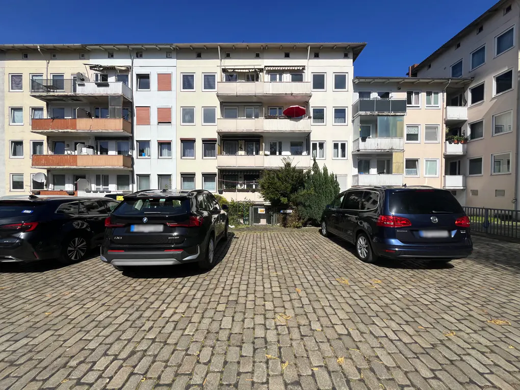 Apartment building with parked cars. The building is light colored with balconies. The parking area is paved with cobblestones.