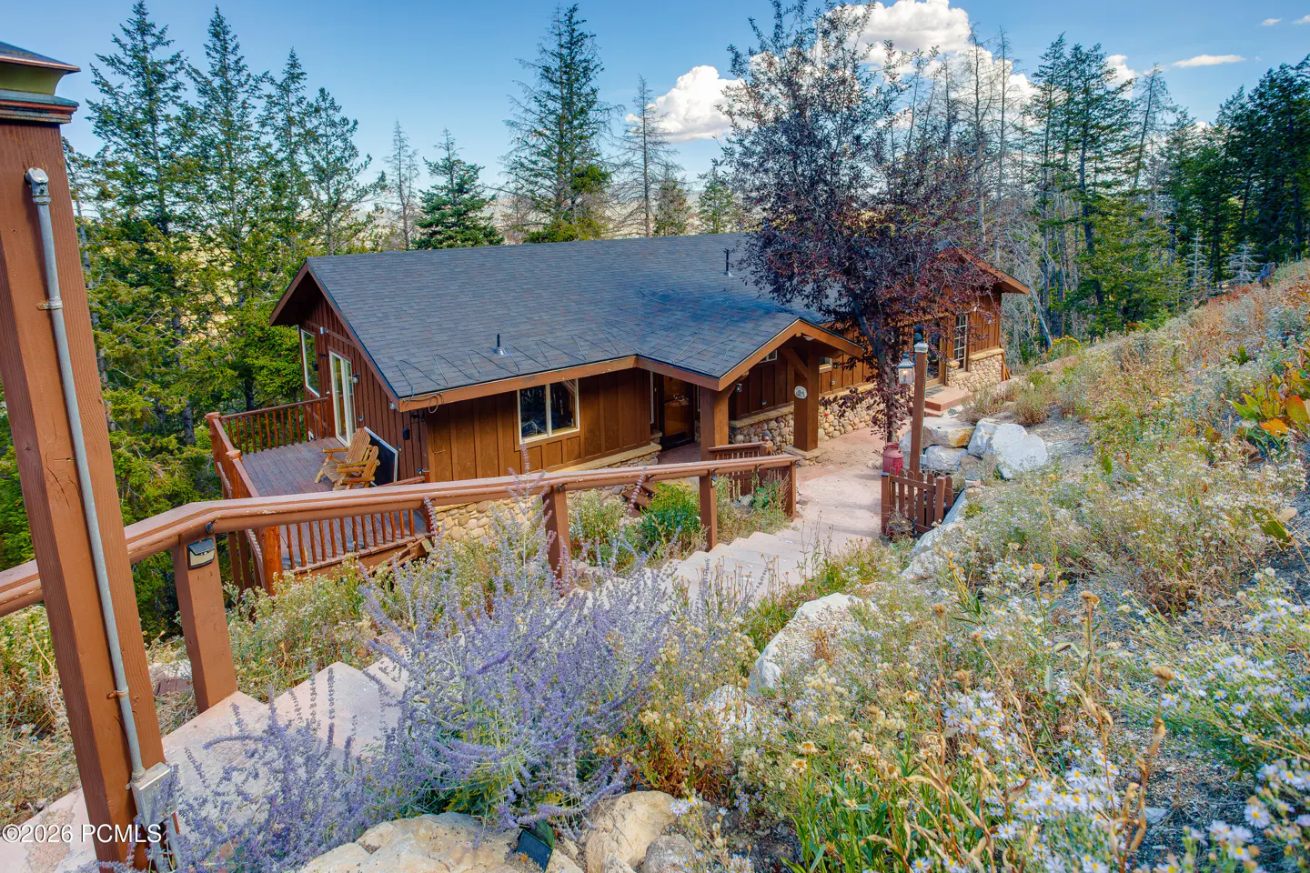 Exterior view of a brown house with a dark roof, surrounded by trees and a hillside with flowers. A wooden staircase leads up to the house.