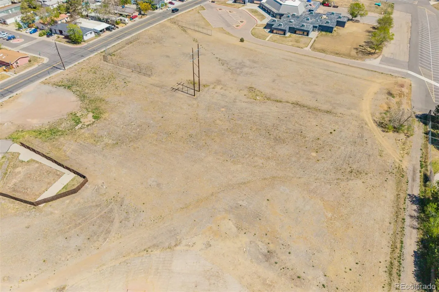 Aerial view of a large, vacant lot with sparse vegetation, utility poles, and a building with a parking lot in the background.
