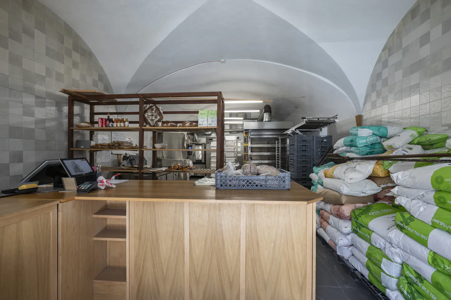 Bakery interior with a wooden counter, shelves with baked goods, and stacks of flour bags. Arched ceiling and tiled walls.
