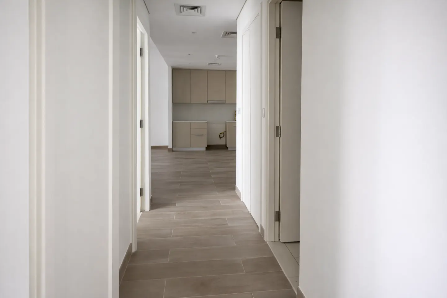 Hallway view of a modern apartment with white walls, light brown tile flooring, and a kitchen in the background with light brown cabinets.