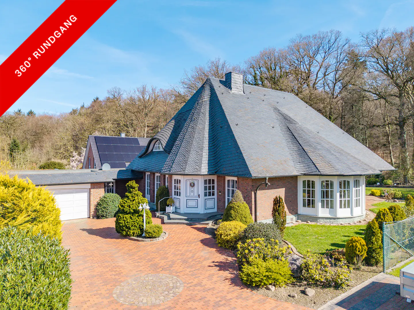 A brick house with a gray roof, white trim, and a brick driveway is surrounded by green bushes and trees. A red banner reads "360° Rundgang."