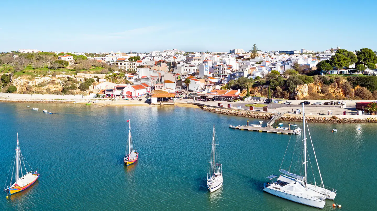Aerial view of Ferragudo, Portugal, with sailboats on the Arade River and white buildings with red roofs on the shore.
