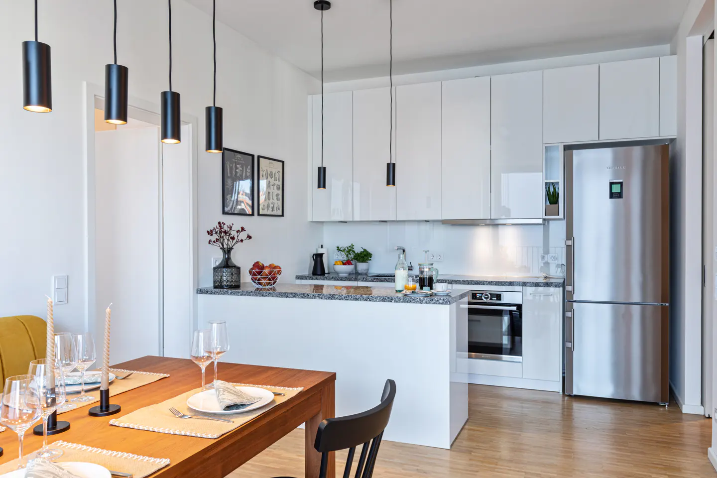 Bright, modern kitchen with white cabinets, stainless steel refrigerator, and granite countertops. A wooden dining table is set with glasses and plates.