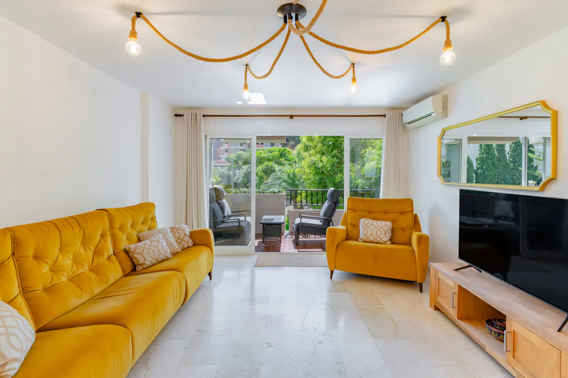 Bright living room with yellow sofa and chair, marble floors, and a balcony with green trees visible through the sliding glass doors. A rope light fixture hangs from the ceiling.