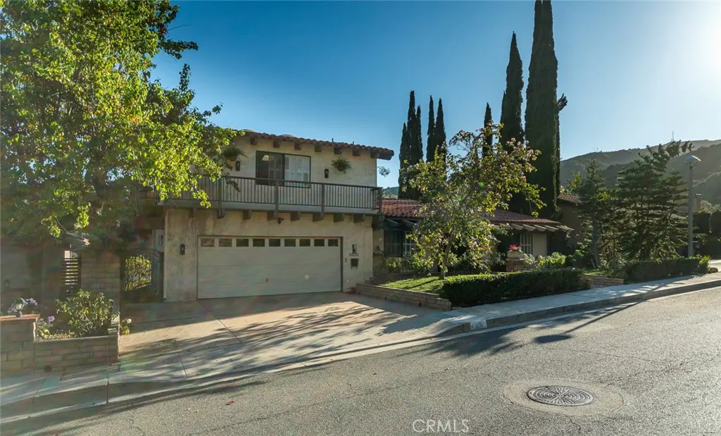 Two-story Spanish-style home with a beige stucco exterior, tile roof, balcony, and garage, set against a sunny hillside.