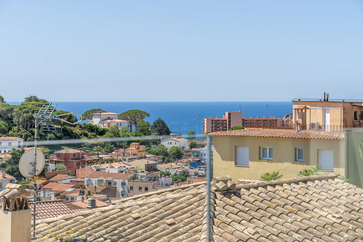 Elevated view of a coastal town with terracotta roofs, lush trees, and a clear blue sea under a bright sky.