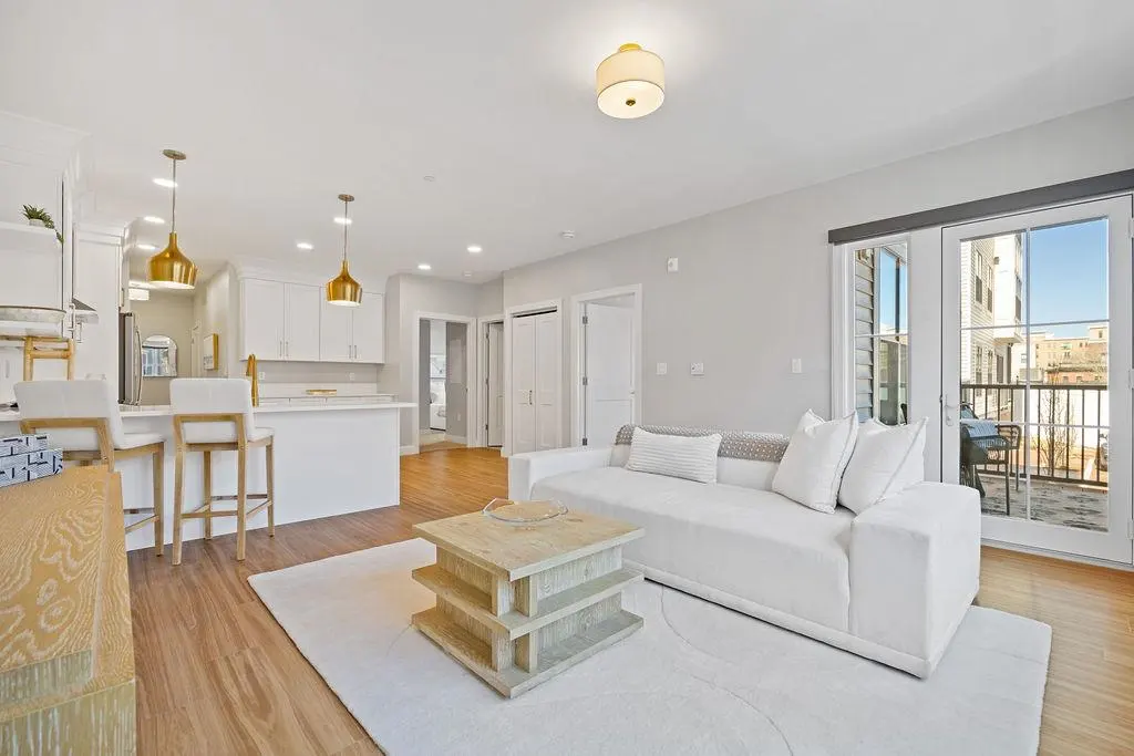 Bright, open-concept living space with white sofa, kitchen island with bar stools, and balcony access. Wood floors and neutral tones.