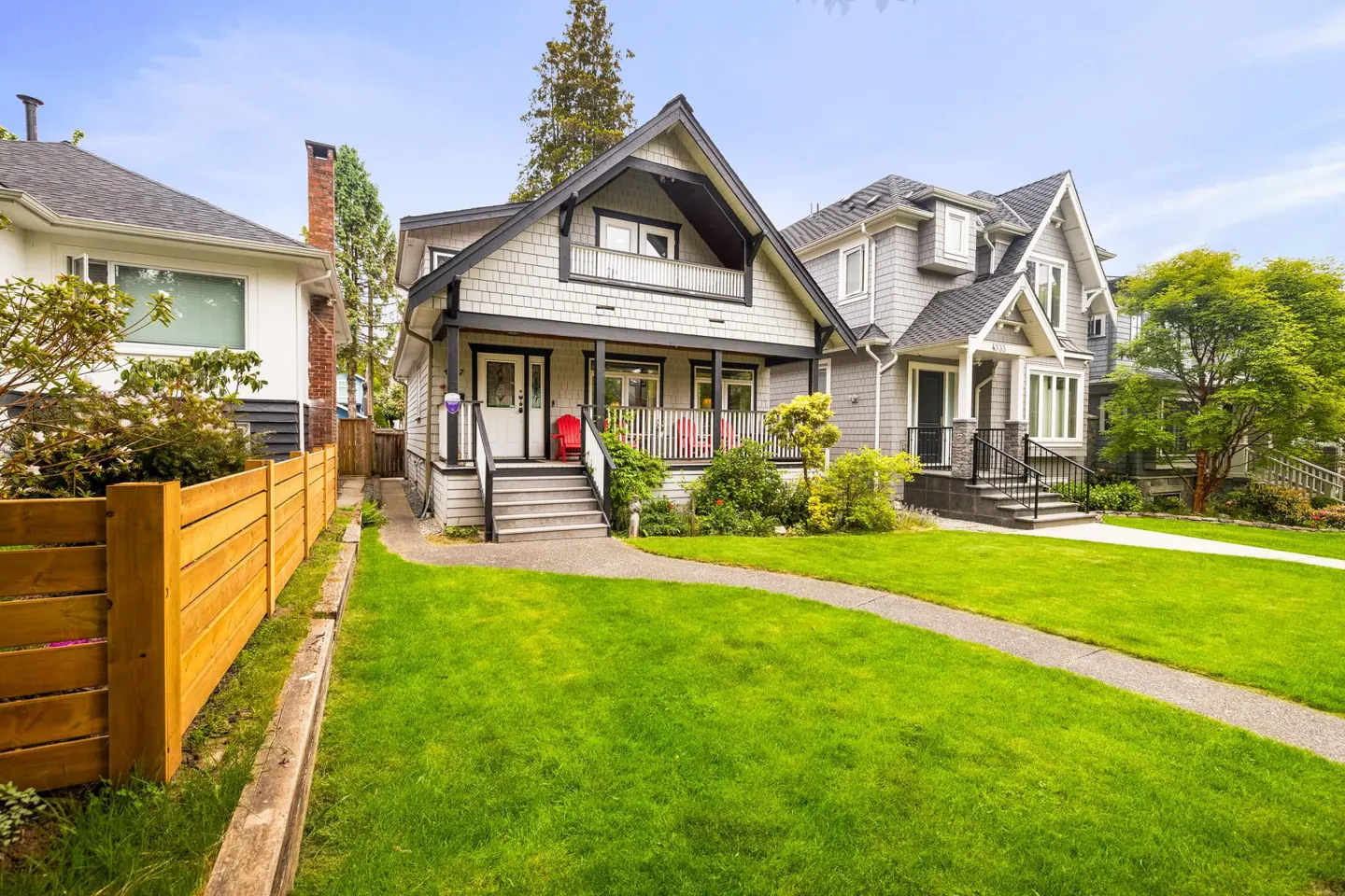 A gray two-story house with a green lawn and a wooden fence. Two red chairs are on the porch.