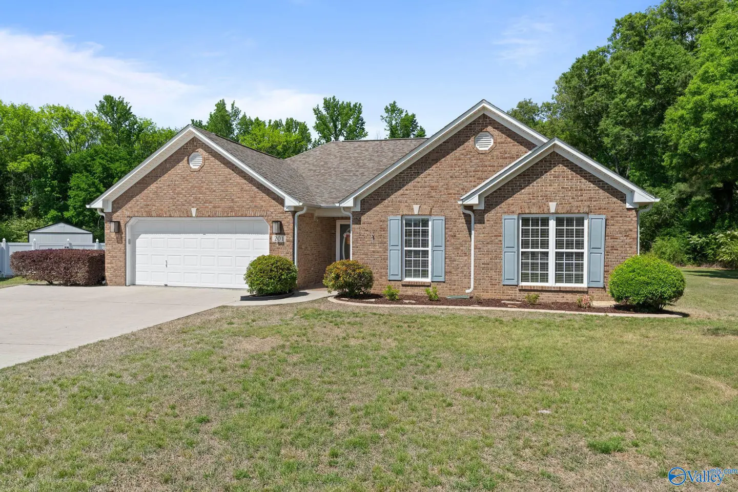 A single-story brick house with light blue shutters and a white garage door. Green lawn and trees in the background.