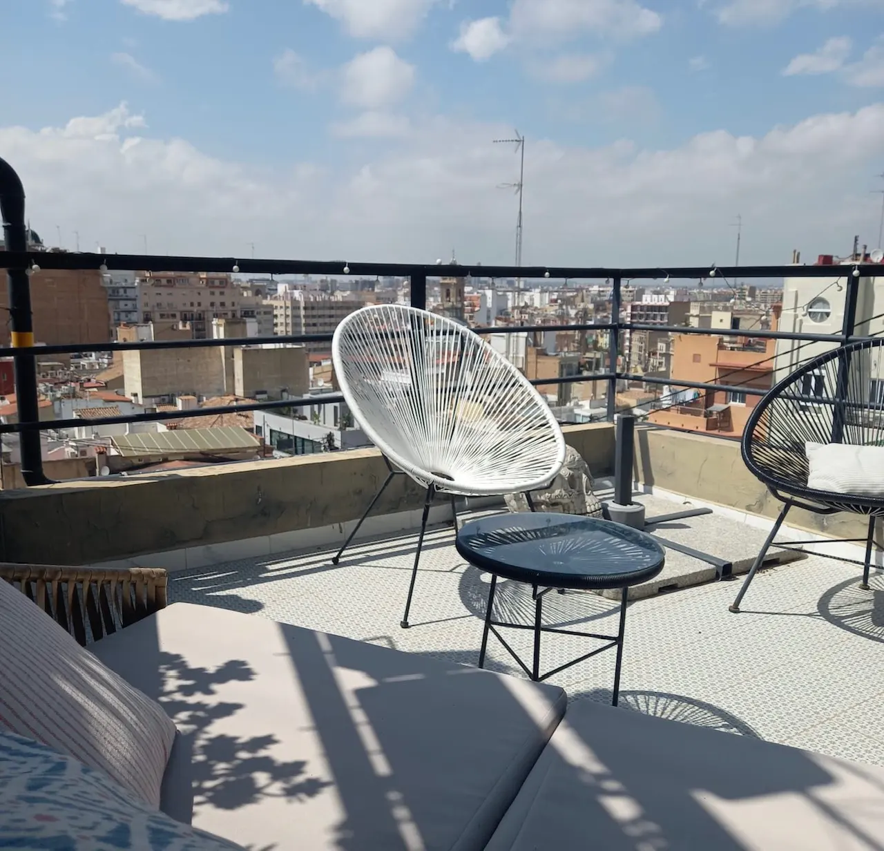 Rooftop patio with white Acapulco chair, black table, and lounge seating. Cityscape view with blue sky and clouds in the background.