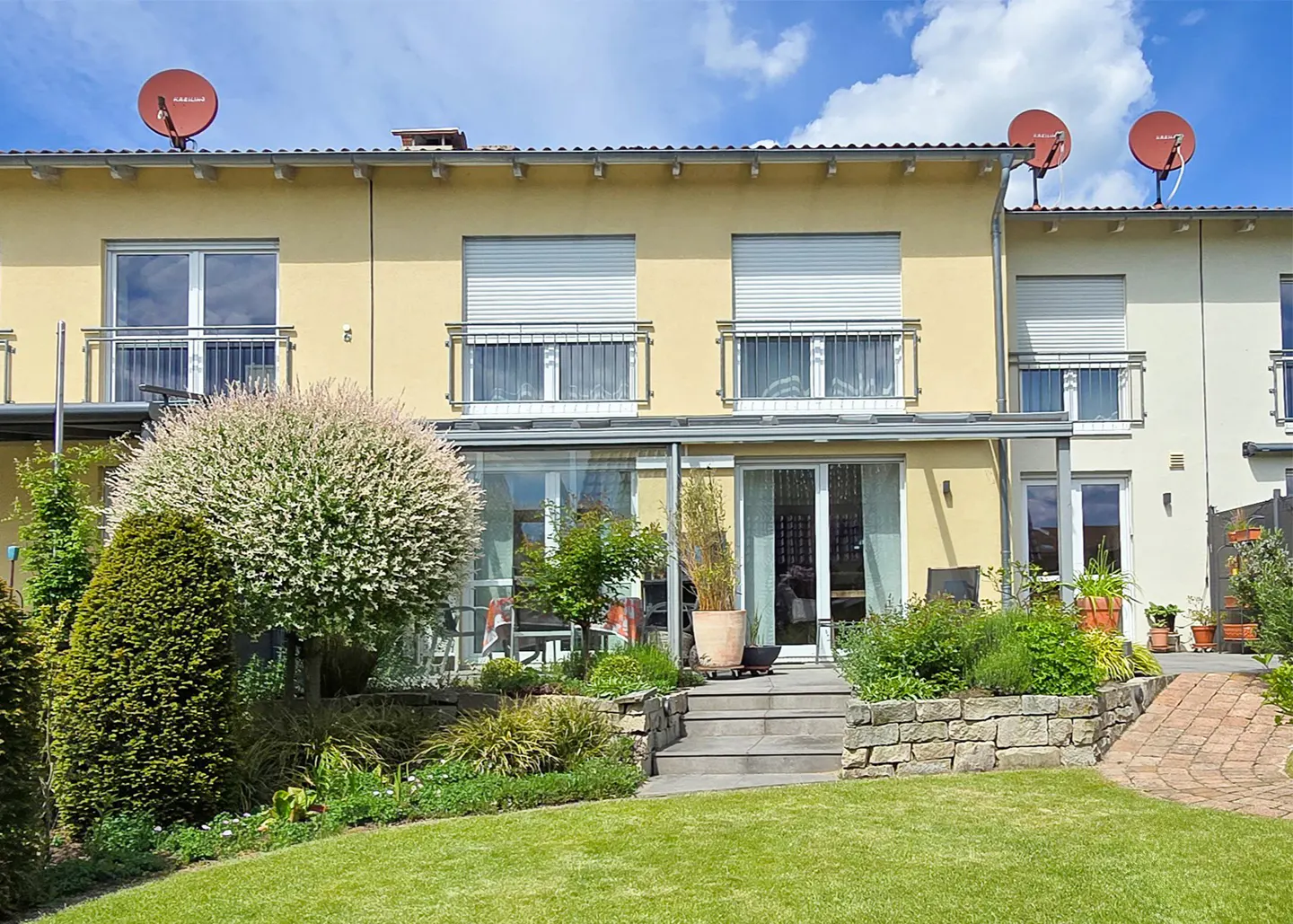 Exterior view of a yellow townhouse with a patio, green lawn, and landscaping. Satellite dishes are on the roof.