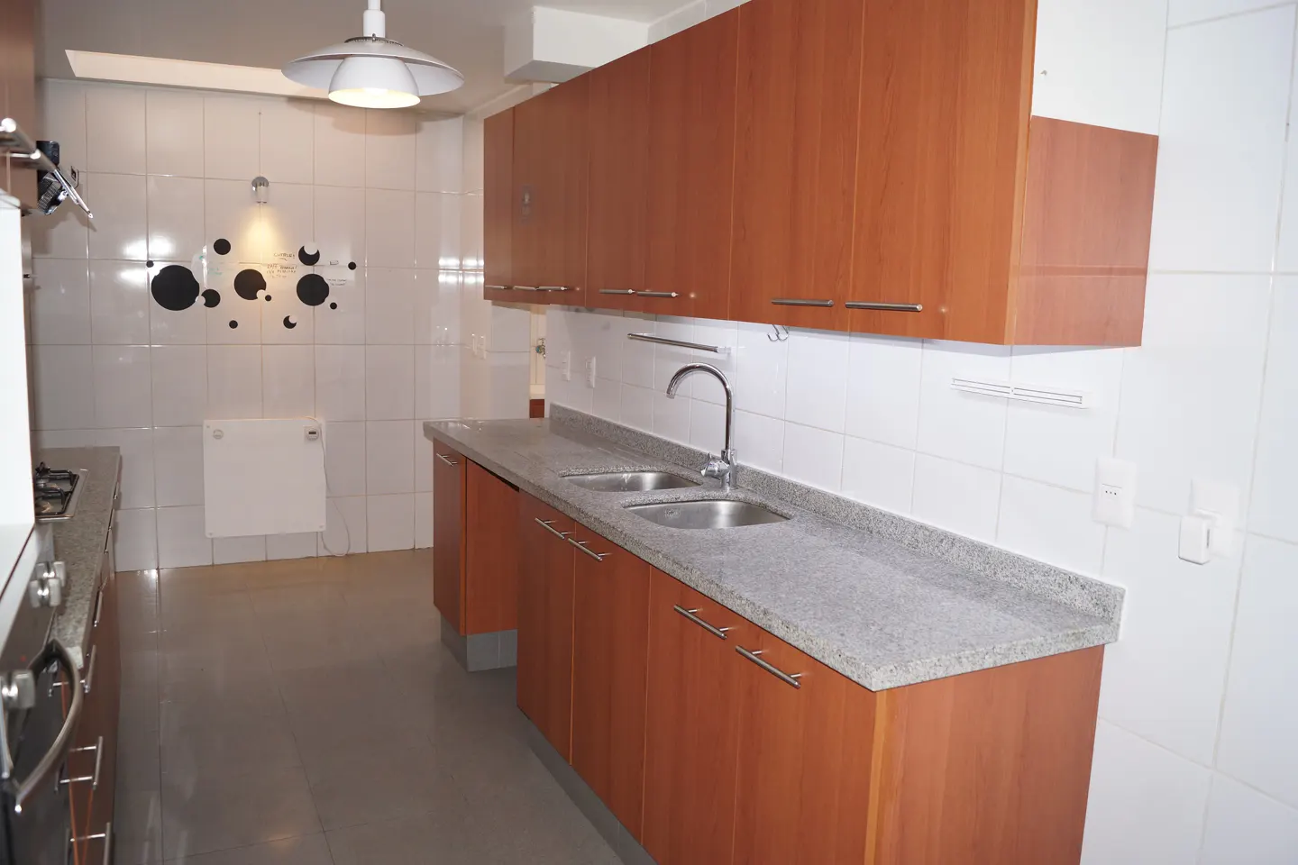 A bright kitchen with wood cabinets, a gray countertop, and white tiled walls. A double sink is visible.