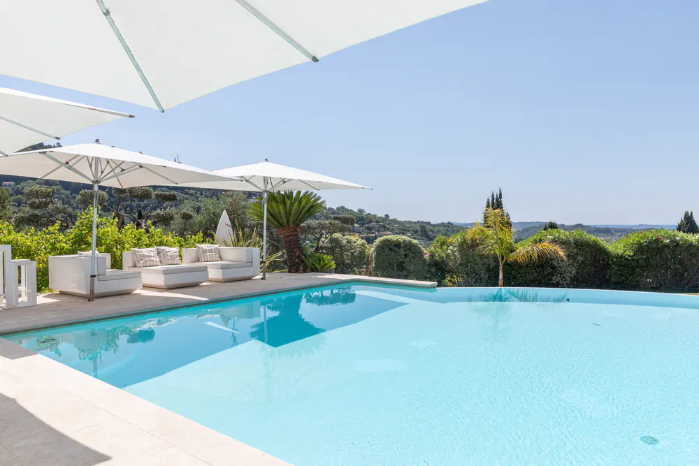 A bright blue swimming pool with white lounge furniture and umbrellas on a patio overlooking a lush, sunny hillside.