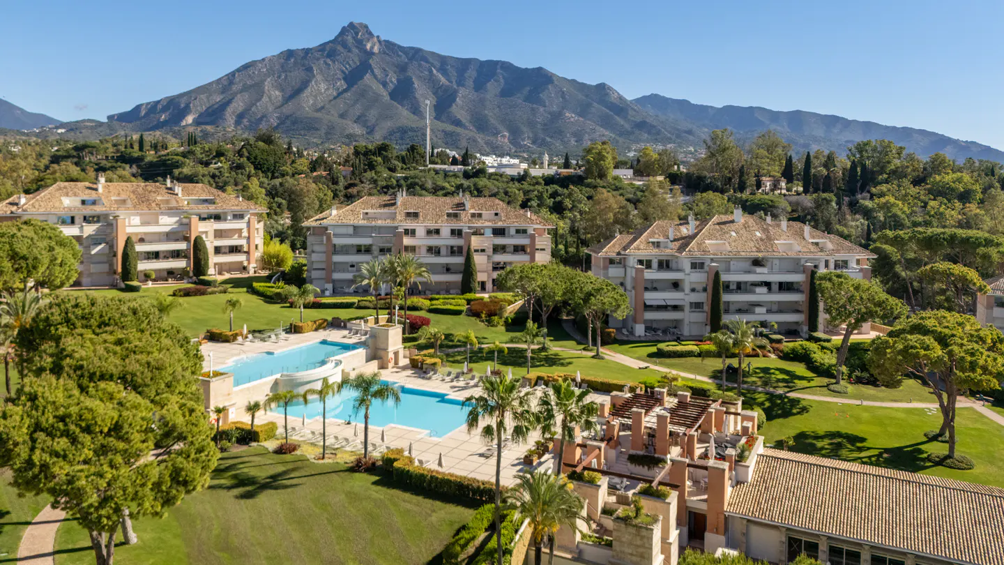 Aerial view of luxury apartments with a pool, green lawns, palm trees, and a mountain backdrop under a clear blue sky.