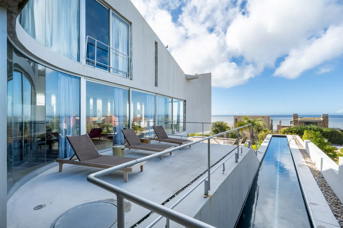 Exterior view of a modern white house with a patio, lounge chairs, and a narrow pool overlooking the ocean.