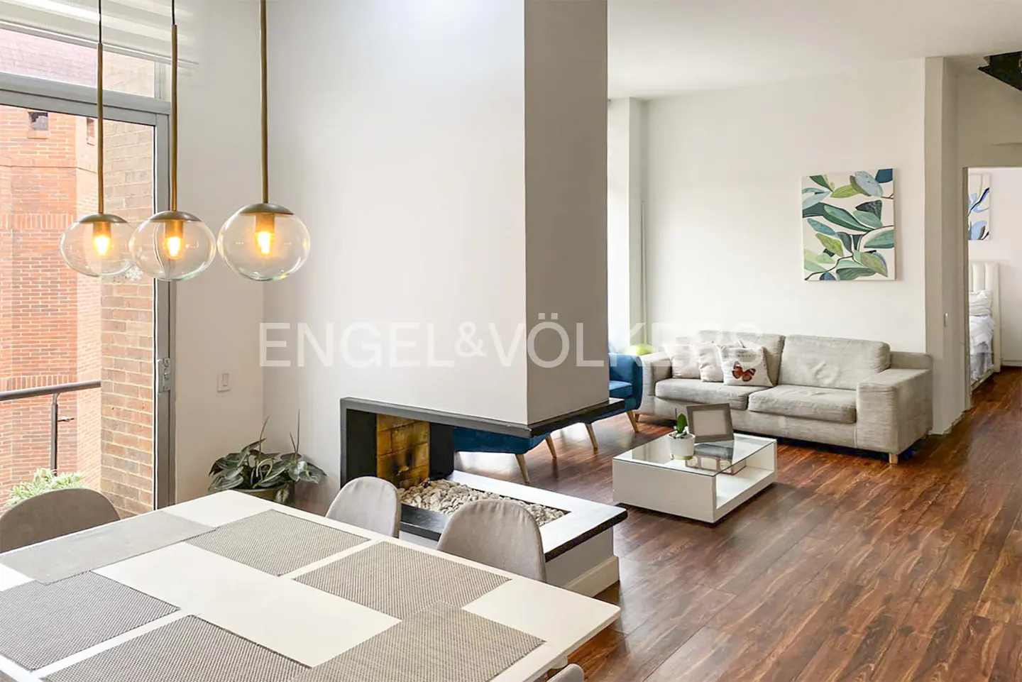 Bright, modern apartment interior with a dining table, fireplace, and living room. Three pendant lights hang above the table.