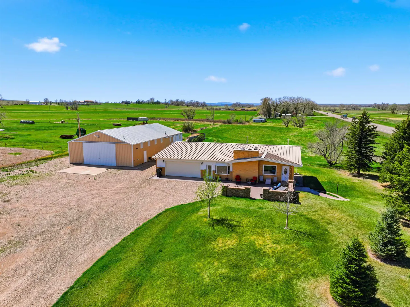 Aerial view of a tan ranch-style home and large metal workshop on a rural property with green fields under a blue sky.