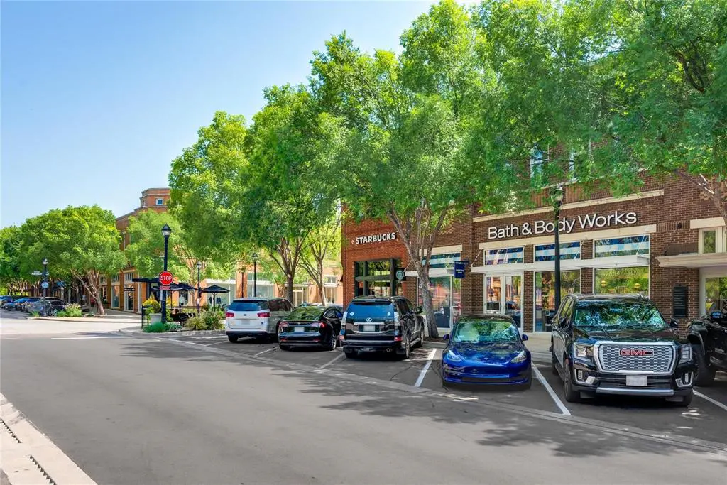 Street view of a shopping center with Starbucks and Bath & Body Works. Cars are parked along the street, shaded by green trees.