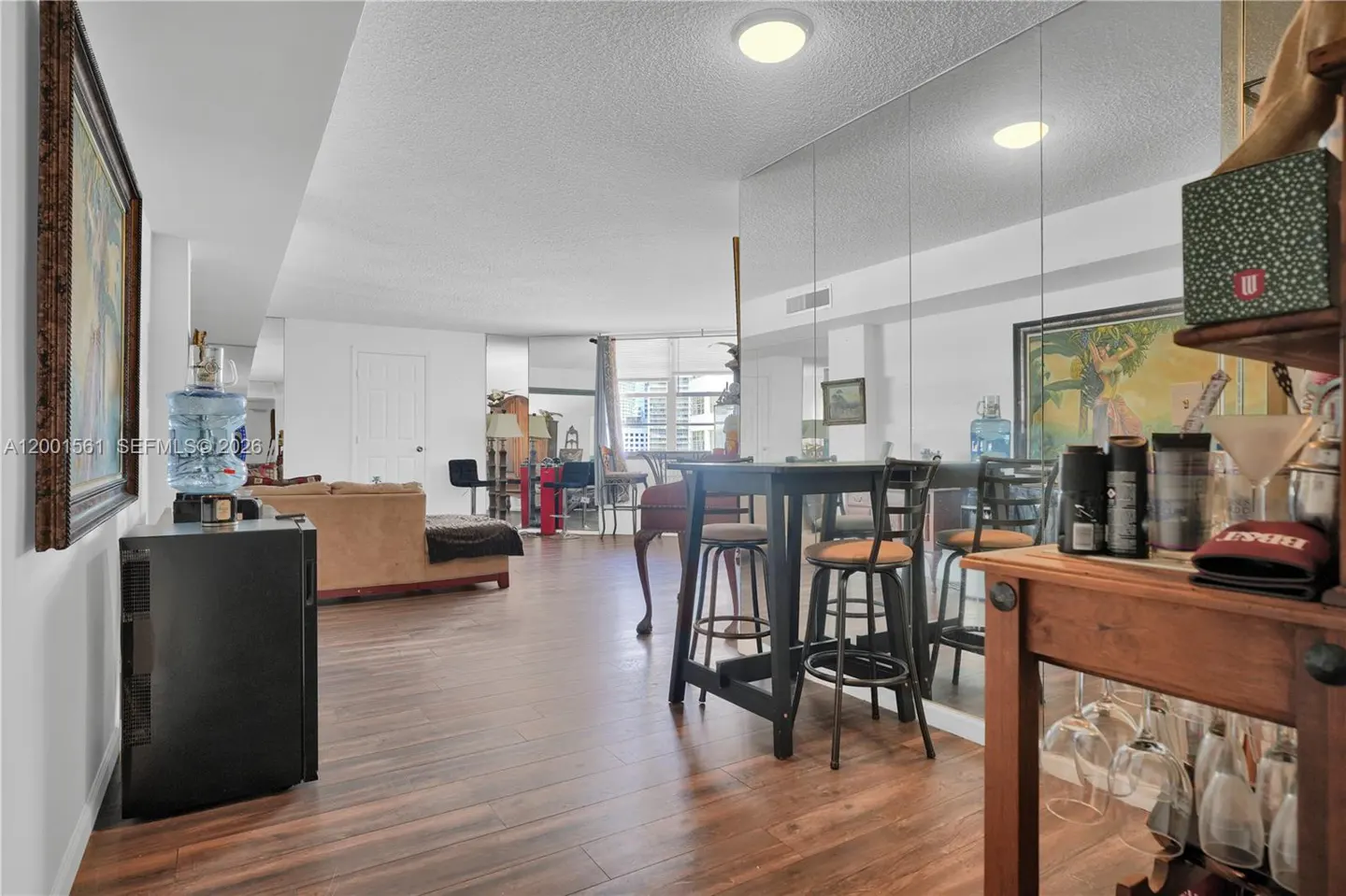 A condo interior with wood floors, a black table with stools, and a mirrored wall. A tan sofa and a painting are visible in the background.