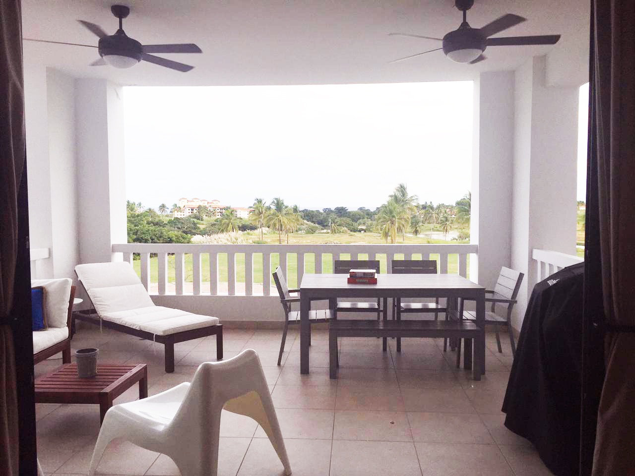 A bright, airy balcony with a table, chairs, and lounge chair overlooks a green landscape with palm trees. Two ceiling fans are visible.