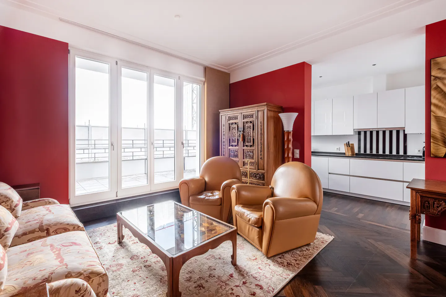 Bright living room with red walls, dark wood floors, and white kitchen. Two tan leather chairs face a glass coffee table on a patterned rug. A floral sofa sits to the left.