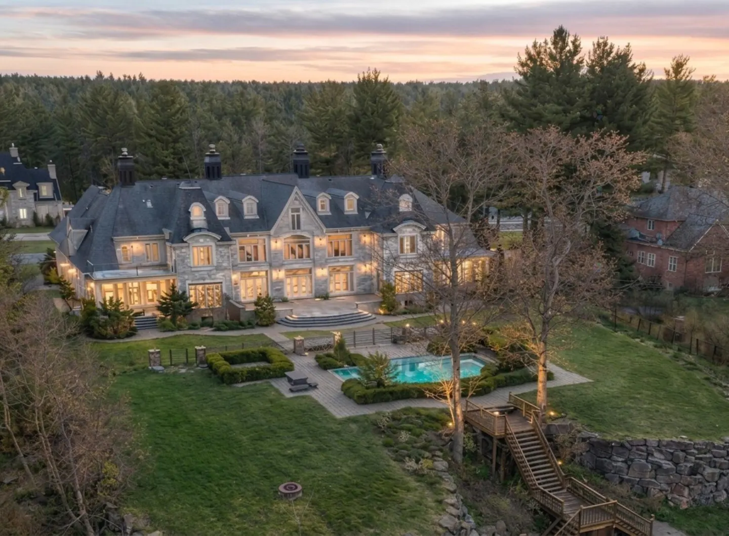 Aerial view of a large, grey stone mansion with a pool, manicured lawn, and forest backdrop at sunset.