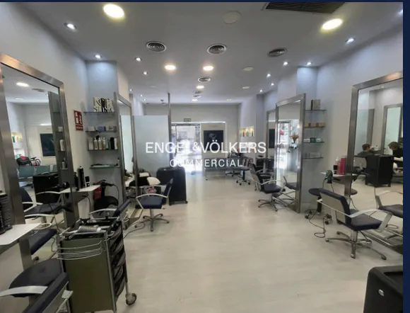 A bright, spacious hair salon with styling stations, large mirrors, and blue chairs. Shelves display hair products. The Engel & Völkers logo is visible.