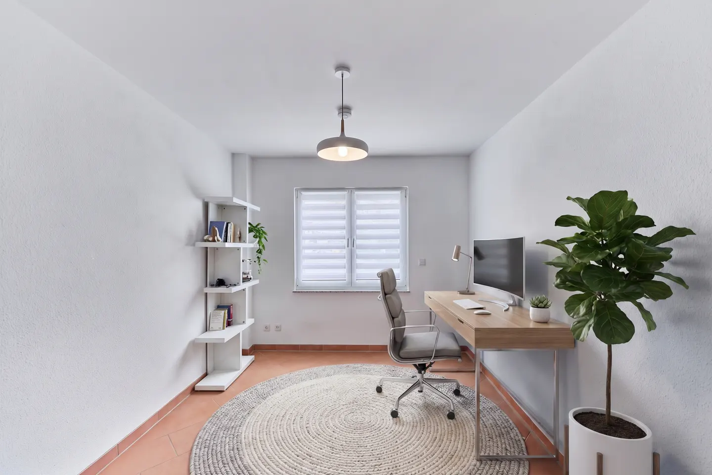 Bright home office with white walls, a desk with a computer, a gray chair, and a large potted plant. A round rug sits on the terracotta floor.