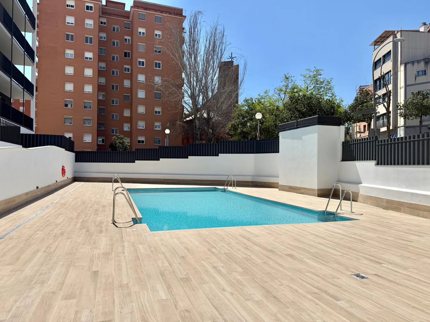 Outdoor pool with turquoise water and metal ladders, surrounded by light wood-look tile. Tall brick buildings in the background.