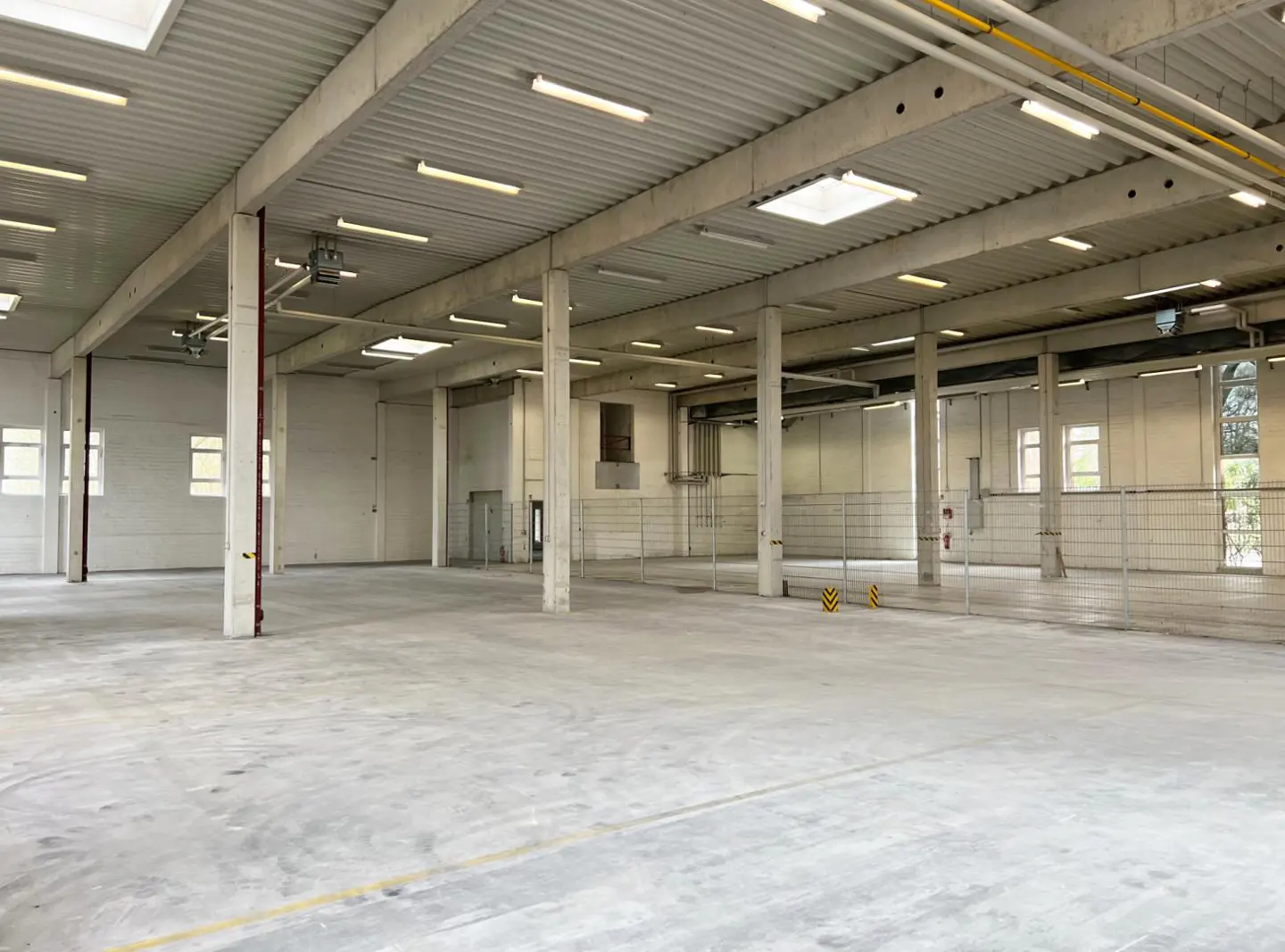 Empty warehouse interior with concrete floor, white walls, and high ceiling with fluorescent lights. Metal safety fence in the background.