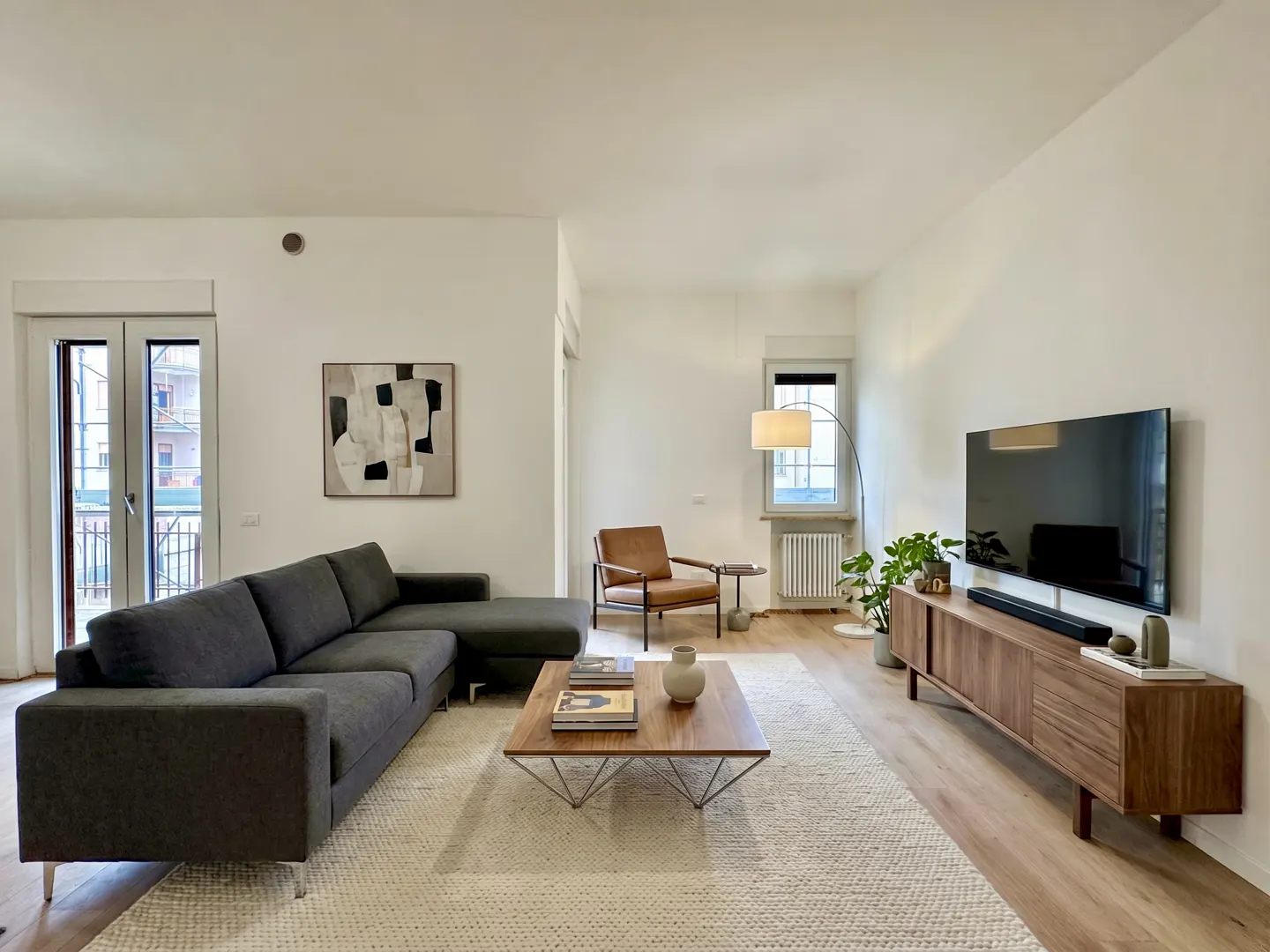 Bright living room with gray sectional sofa, wood coffee table, and TV on a wood console. A tan leather chair sits near a window.