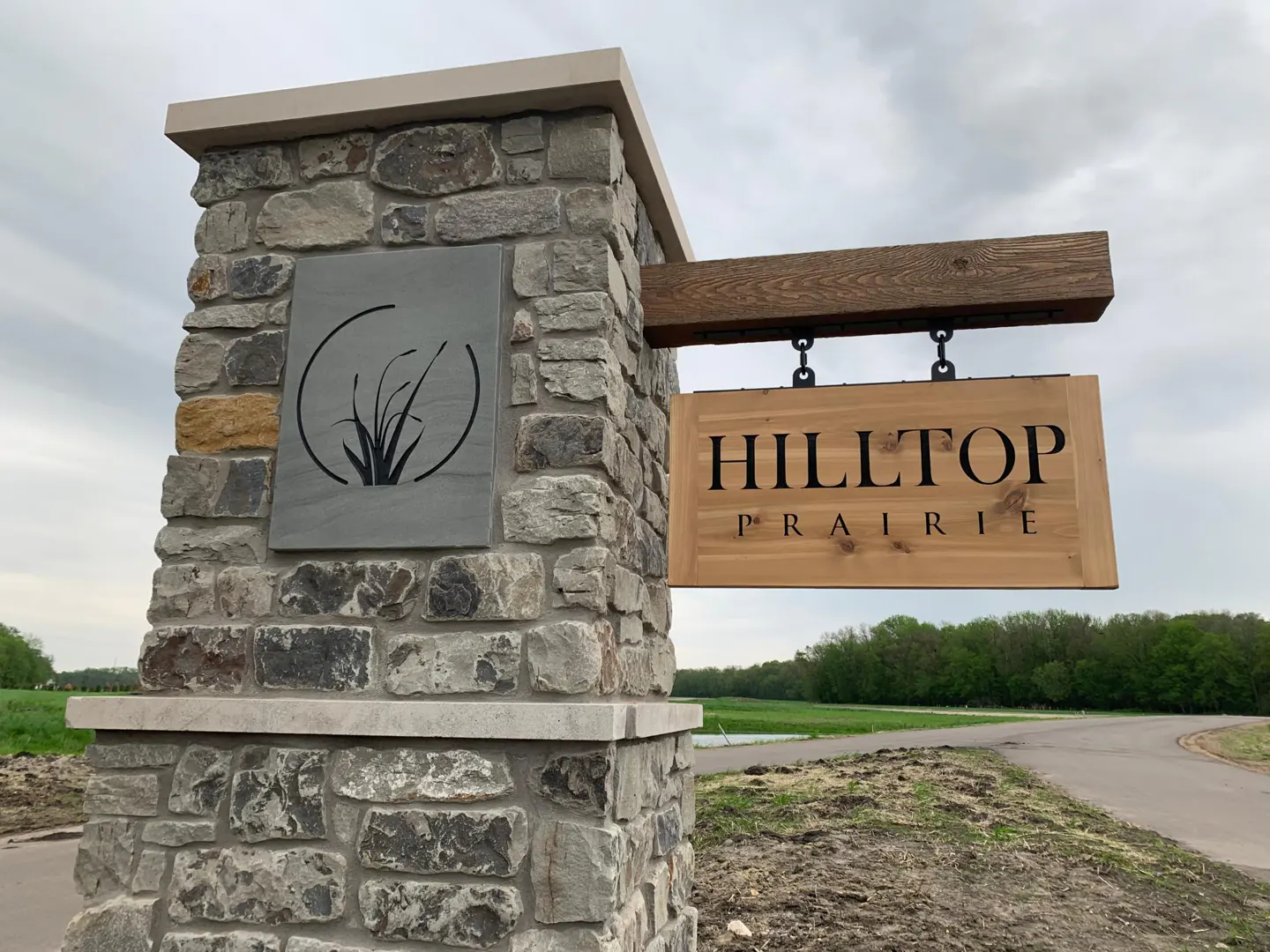 Stone pillar with "Hilltop Prairie" sign. The sign is wood with black lettering, hanging from a wooden beam. A gray stone with a grass design is on the pillar.