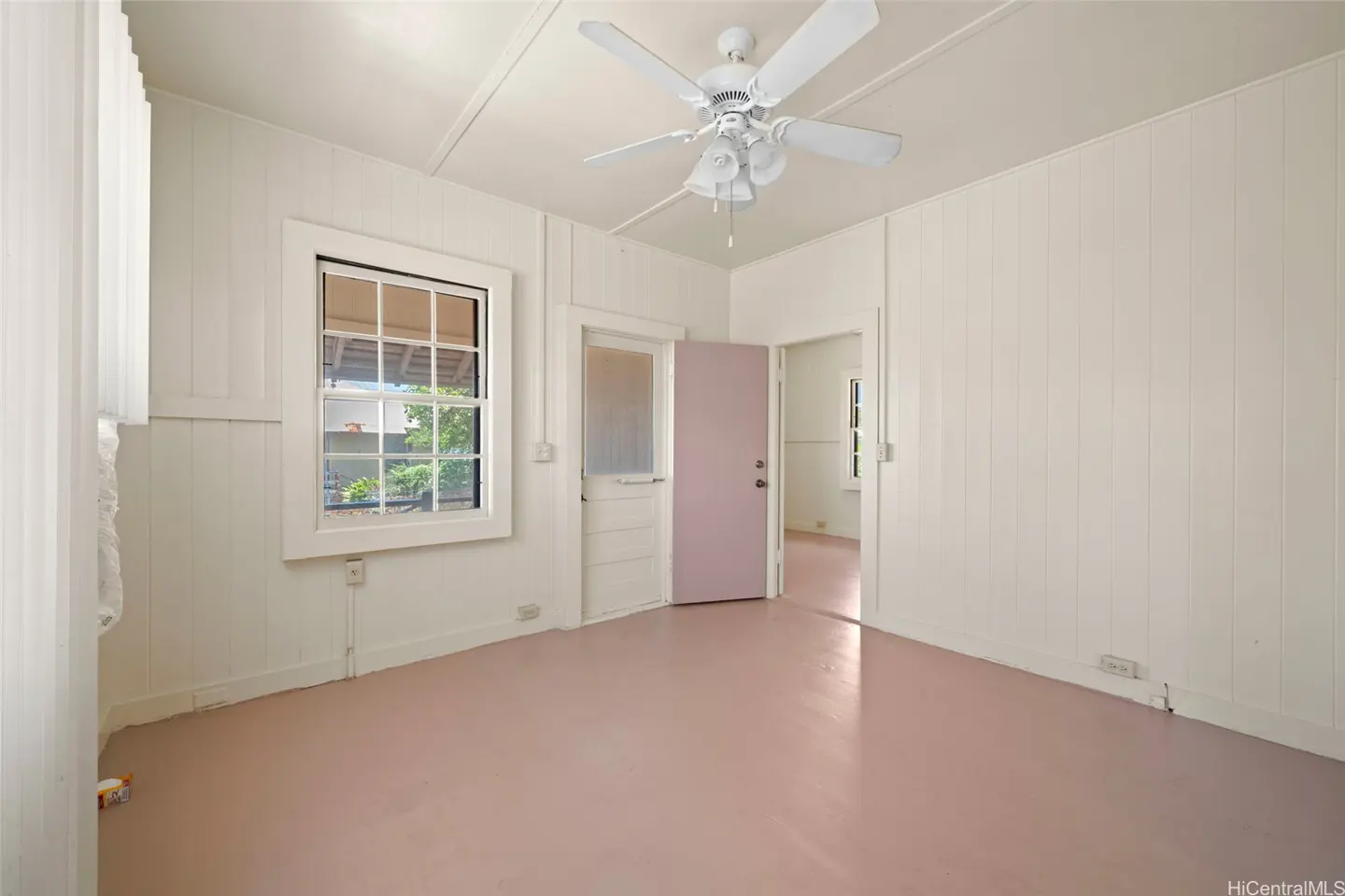 A bright, empty room with white walls, a pink floor, and a white ceiling fan. A window shows greenery outside.
