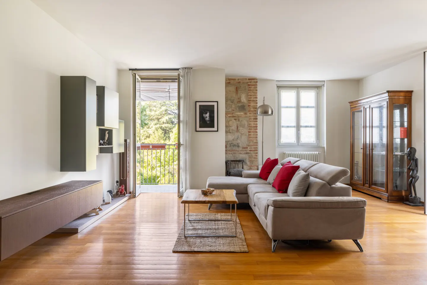 Bright living room with hardwood floors, a gray sectional sofa with red pillows, and a balcony with green trees.