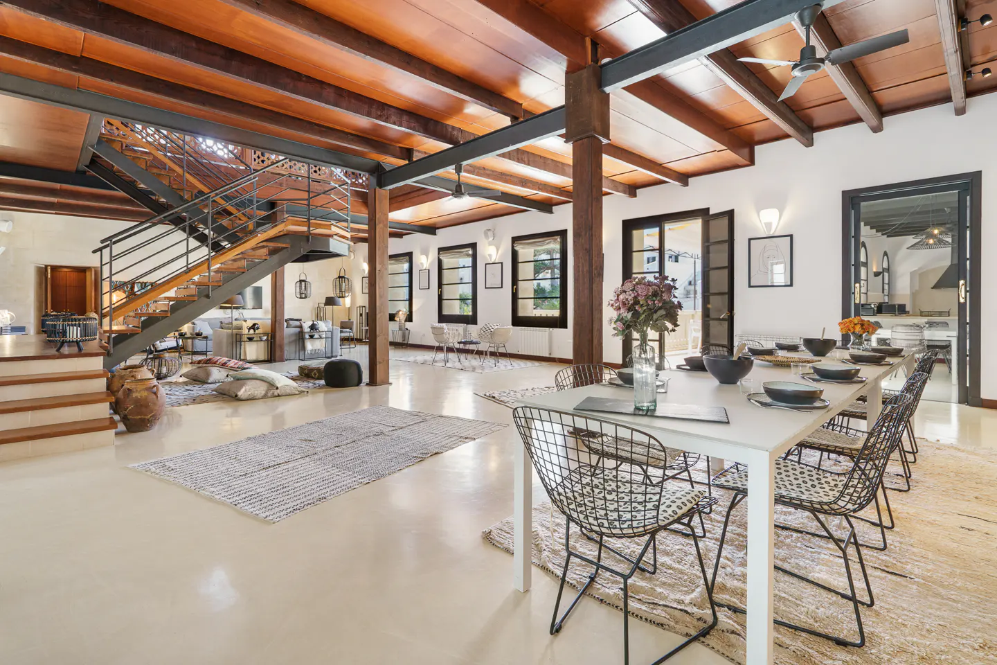 Open-plan living space with a long white dining table, black wire chairs, and wooden beams. A staircase leads to a second floor.
