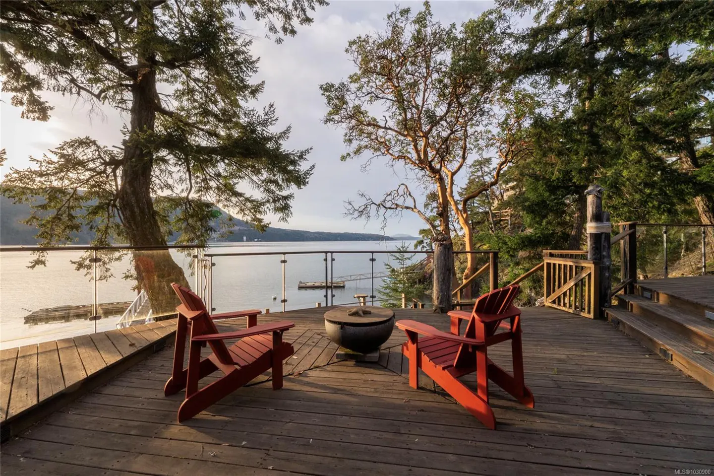 Wooden deck with two red Adirondack chairs facing a fire pit and ocean view, framed by trees.
