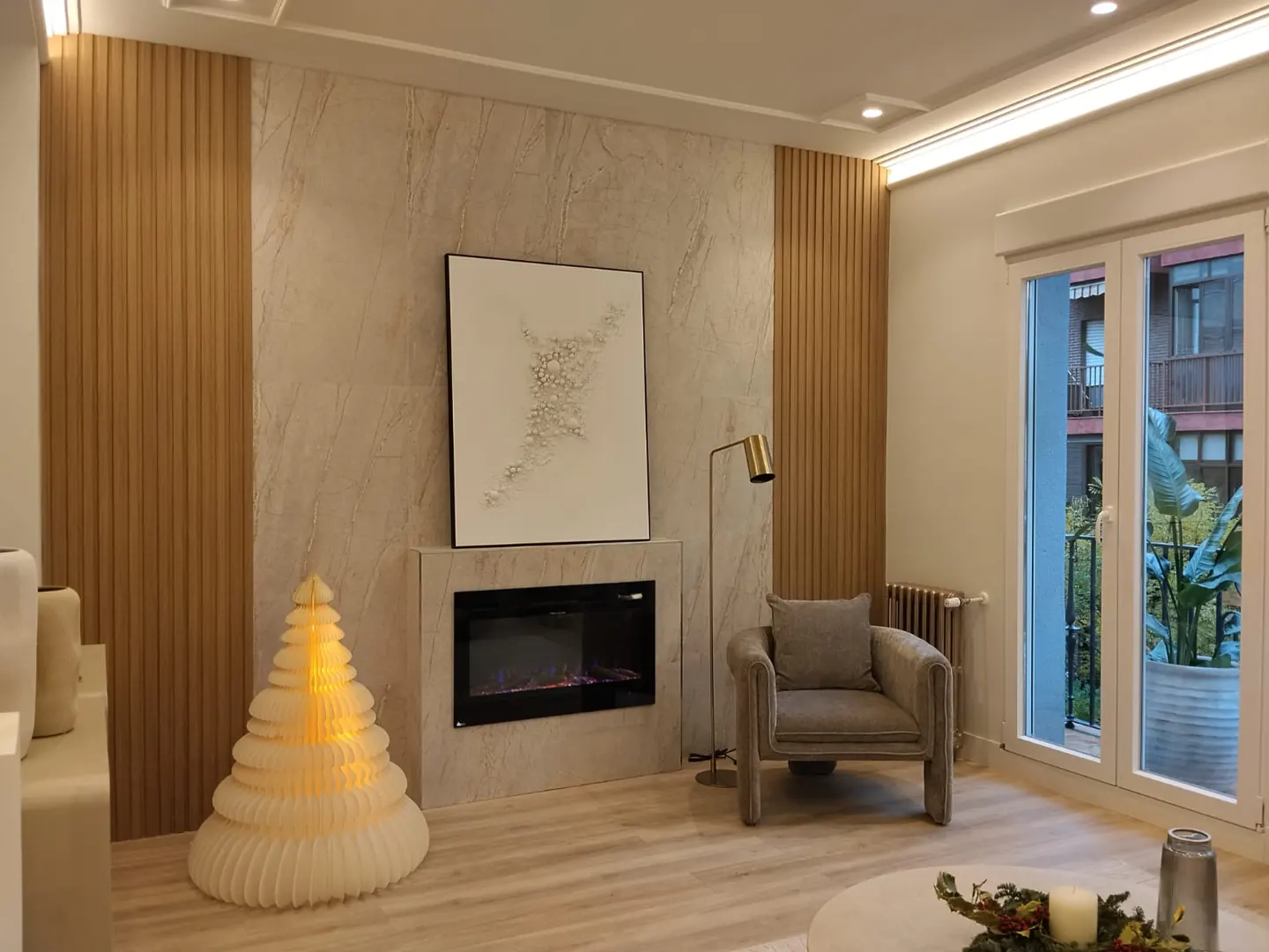 Living room with a fireplace, a gray armchair, and a Christmas tree. A large window overlooks a balcony with plants.
