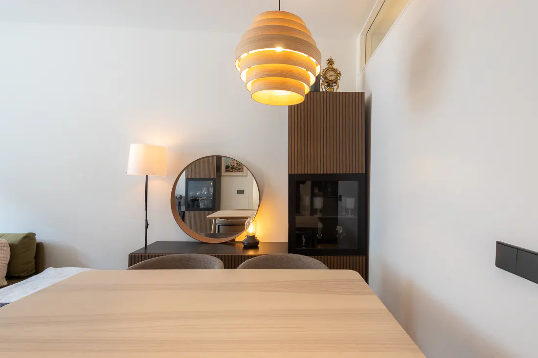 A bright dining room with a light wood table, chairs, and a modern wood slat cabinet with glass doors. A round mirror and lamp sit on a dark wood console.
