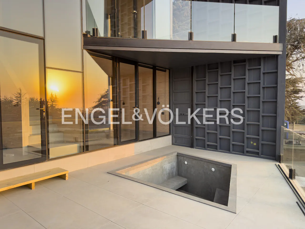 Exterior view of a modern home with glass walls reflecting a sunset, a sunken patio, and the Engel & Völkers logo.