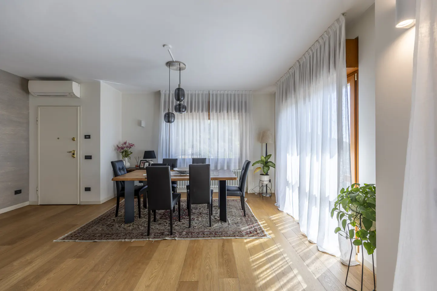 Bright dining room with wood floors, a table with black chairs, and sheer white curtains. A patterned rug sits under the table.