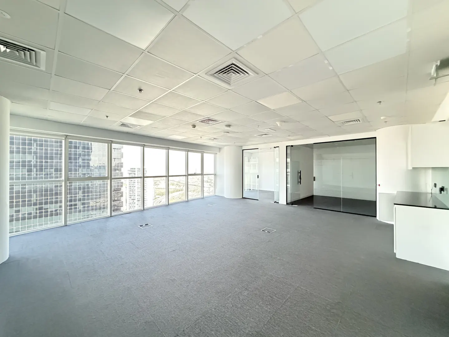 Bright, empty office space with gray carpet, white walls, and large windows with a city view. Glass-walled offices are visible.